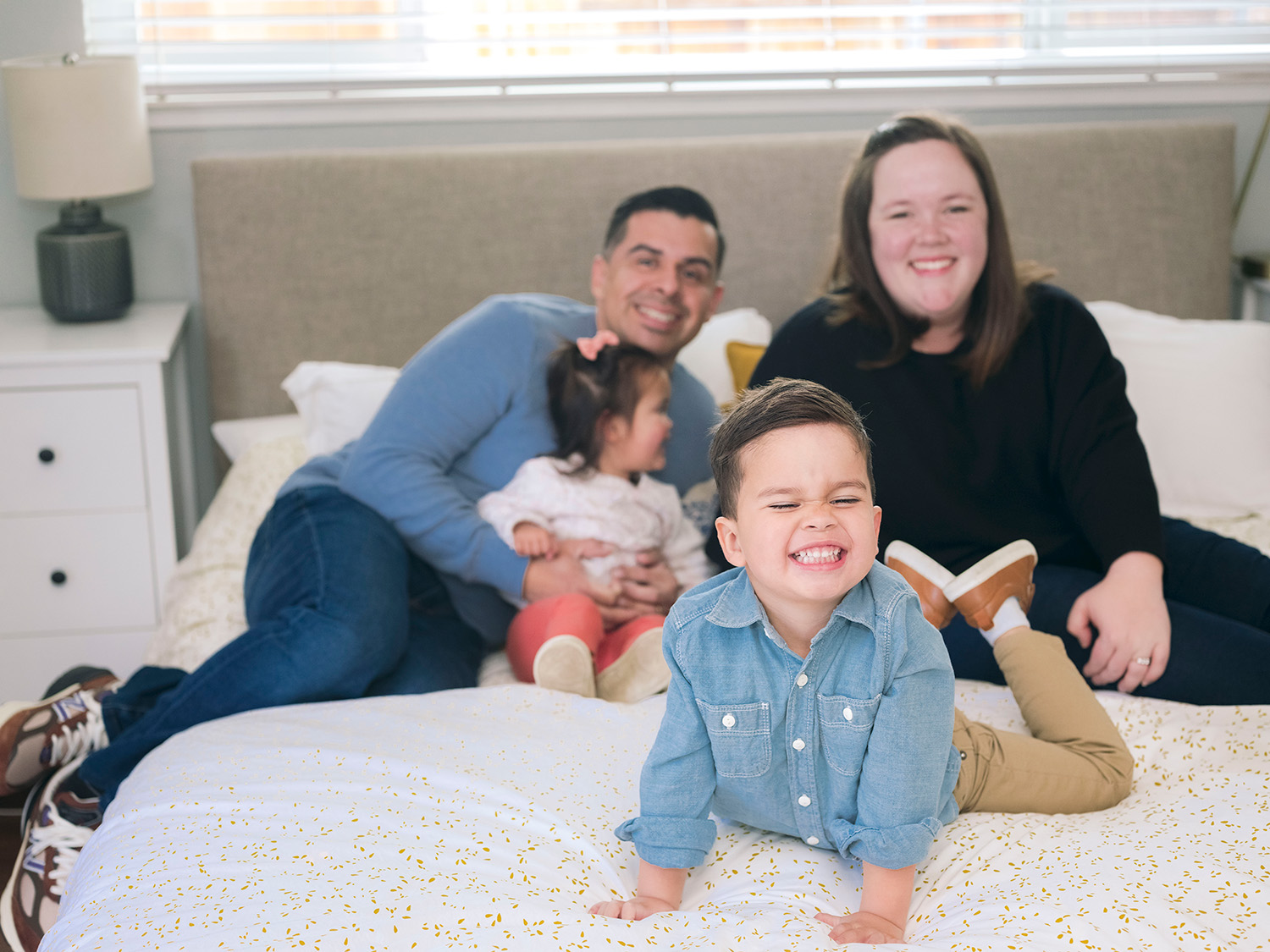 In-home family photo on bed with toddler son being silly and parents laughing in Palo Alto home