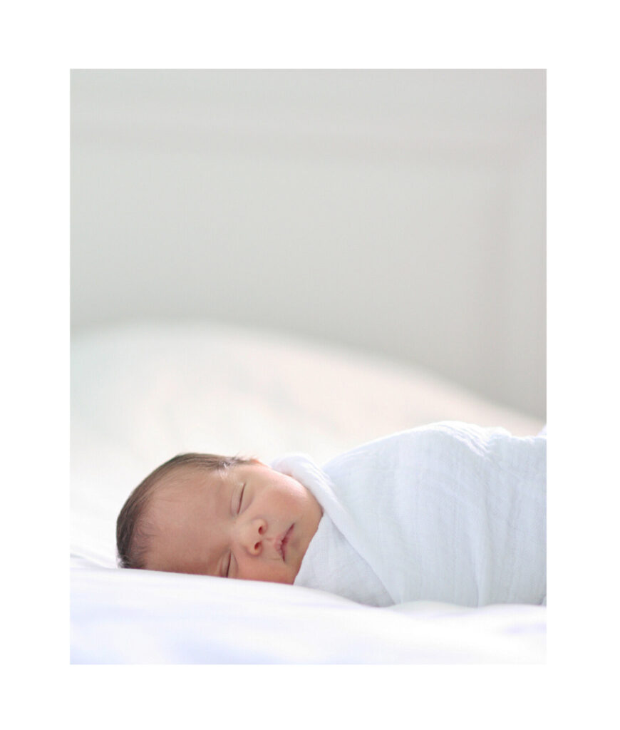 Newborn baby peacefully sleeping on a white bed, wrapped in a soft white blanket during an in-home photo session in Palo Alto by natural newborn photographer Isela Lozano.