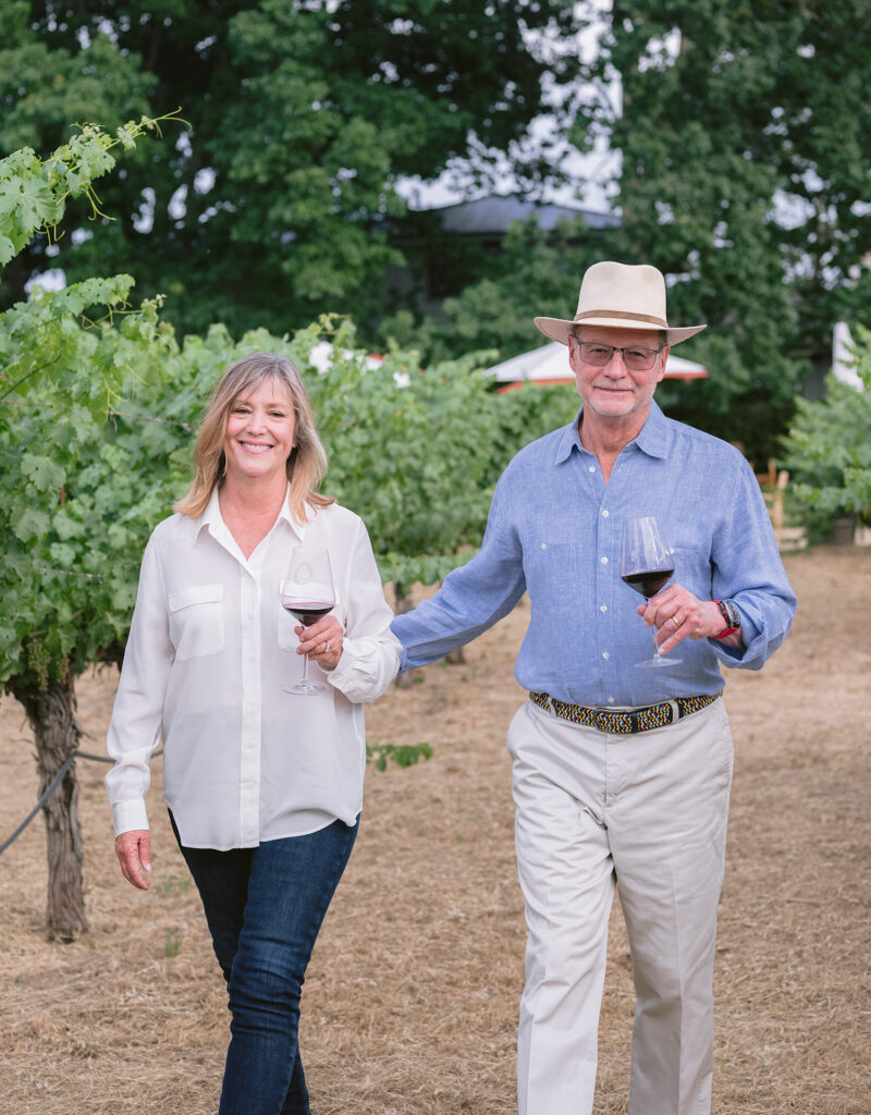 Winery owners walking through vineyard at O'Brien Estate in Napa Valley holding wine glasses during natural light branding session — photographed by Napa Valley winery photographer