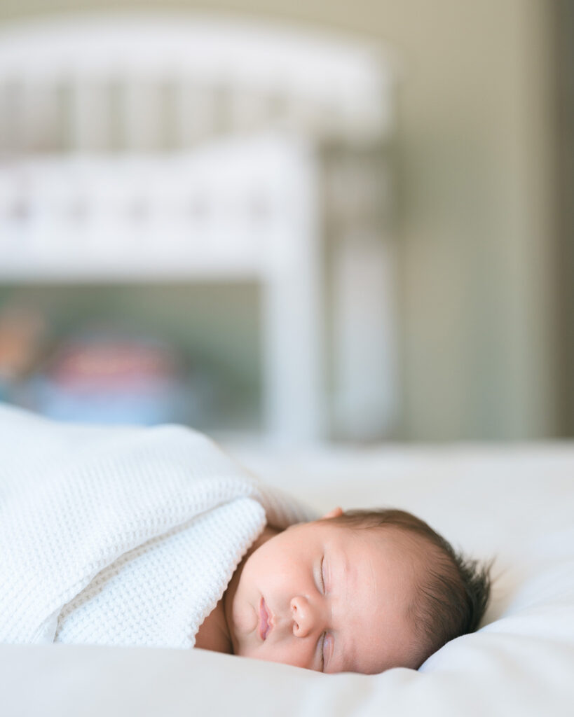 Mill Valley newborn photographer in-home lifestyle session using natural light, newborn baby swaddled in a white blanket resting on a white bed with a crib in the background.