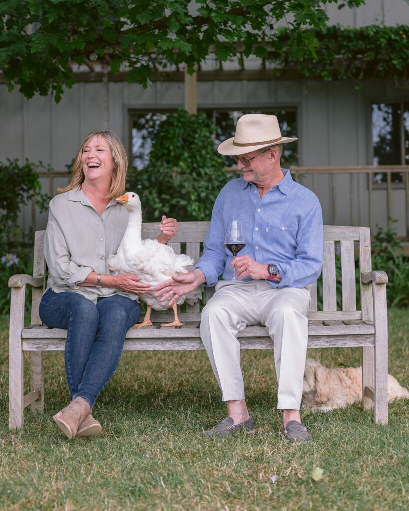 Owners at O'Brien Estate Winery sitting on bench holding a goose while laughing. Captured by Napa Valley Photographer
