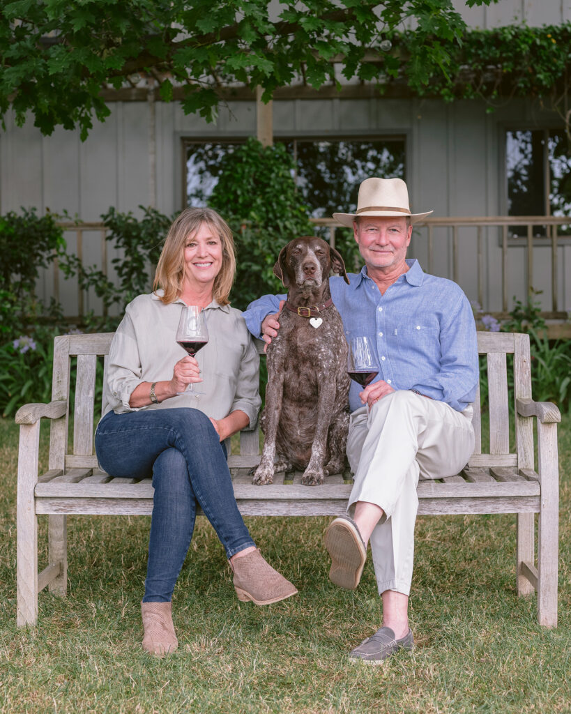 A couple with their dog sitting on a wooden bench outside their Napa Valley winery, holding glasses of red wine during a natural light portrait session by a Napa Valley winery photographer.