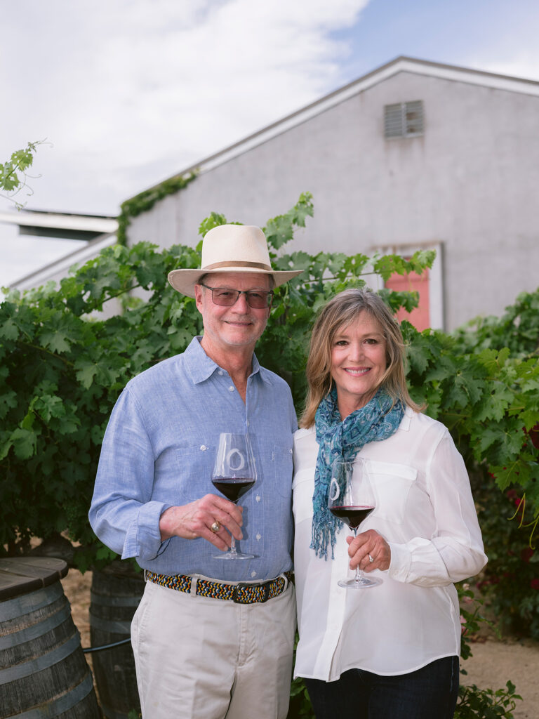Winery owners at O'Brien Estate in Napa standing in the vineyard holding glasses of wine smiling and looking at the camera. Photographed by Napa Valley Photographer