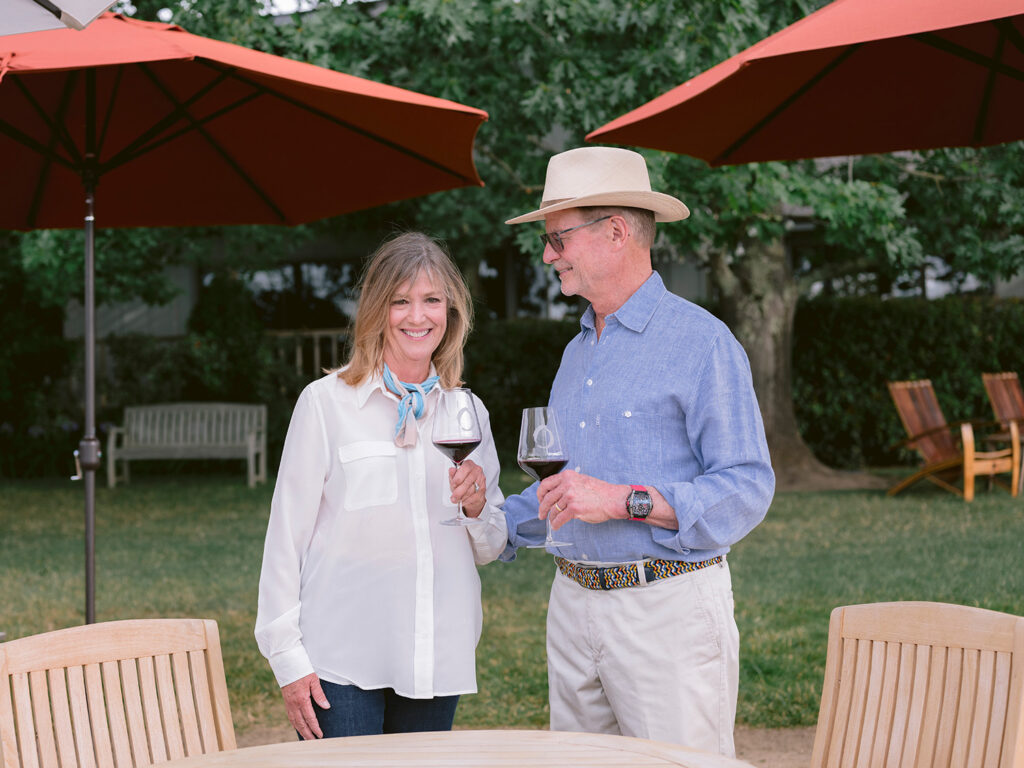 Napa Valley winery owners at O’Brien Estate Winery standing on their vineyard lawn, wife smiling at the camera with a glass of wine while her husband smiles at her, with porch umbrellas and Napa vineyard property in the background.