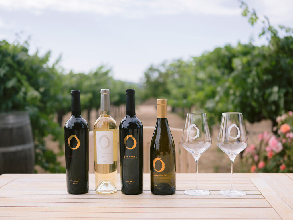 Four wine bottles and two branded wine glasses displayed on a wooden table in a vineyard in natural light at O'Brien Estate in Napa Valley.