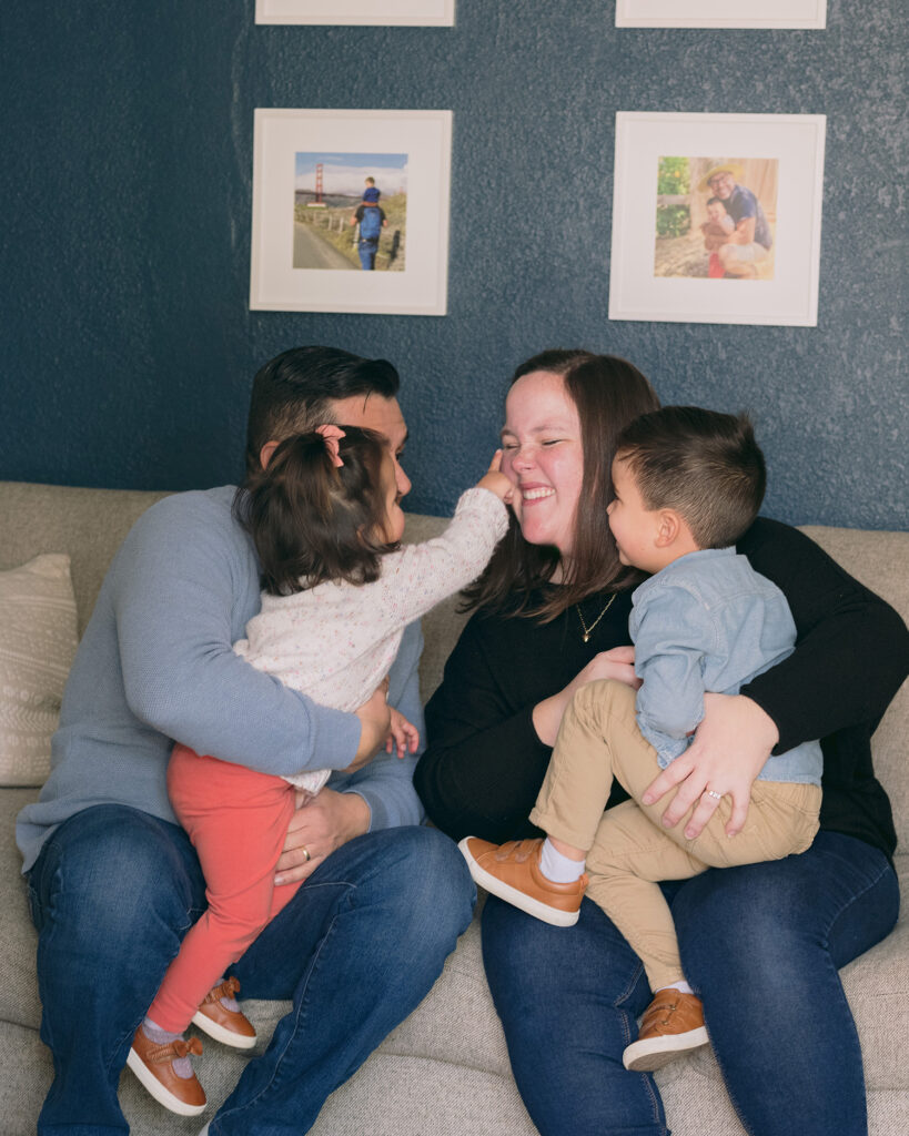 Family of four sitting together on the couch, kids on parents’ laps during an in-home session