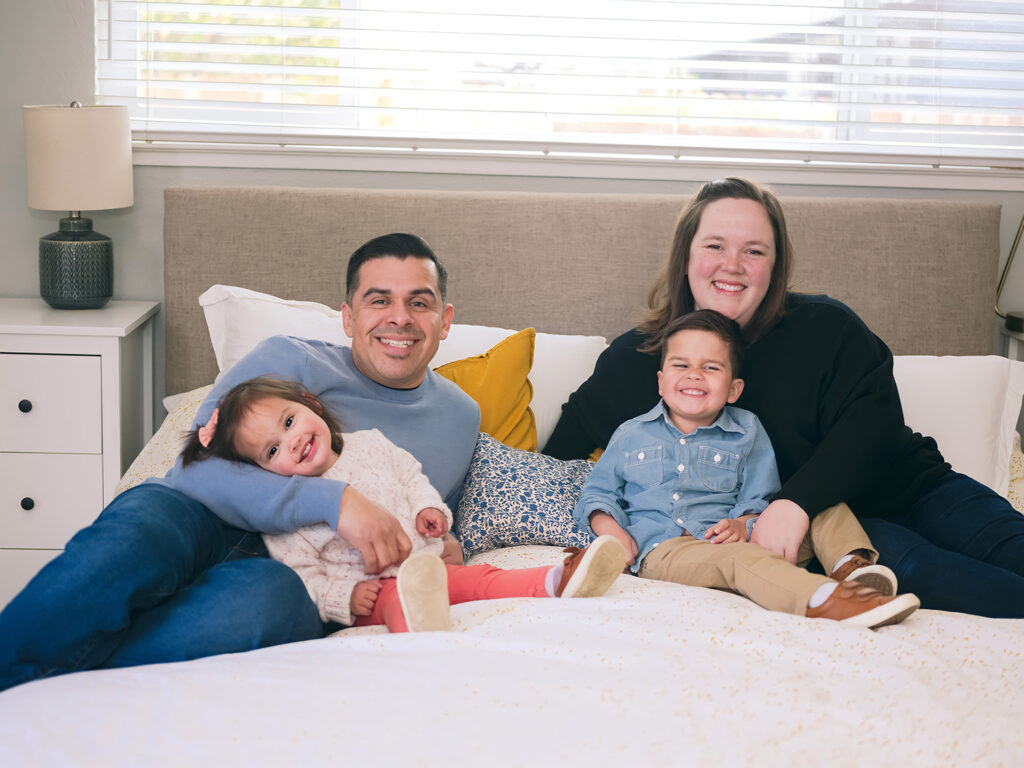 Family of four snuggled together on the parents’ bed, smiling at the camera in soft natural light during their Palo Alto home session