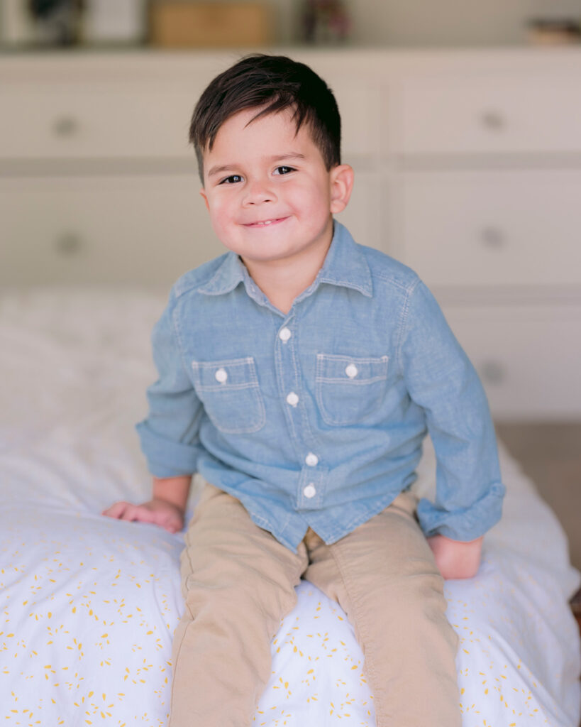 Smiling toddler boy in blue shirt sitting on the edge of a bed in natural light during a lifestyle family session