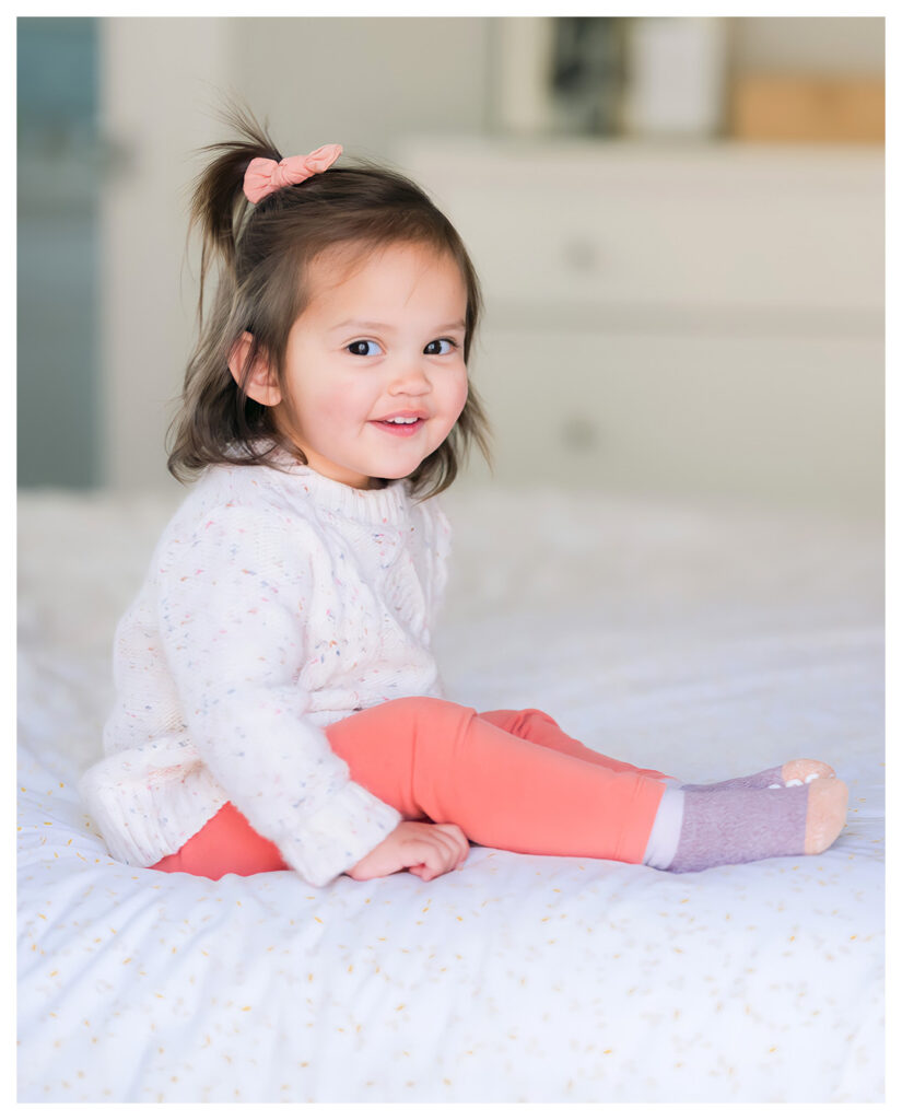 Toddler girl in pink smiling on the bed during a warm, natural light-filled family session