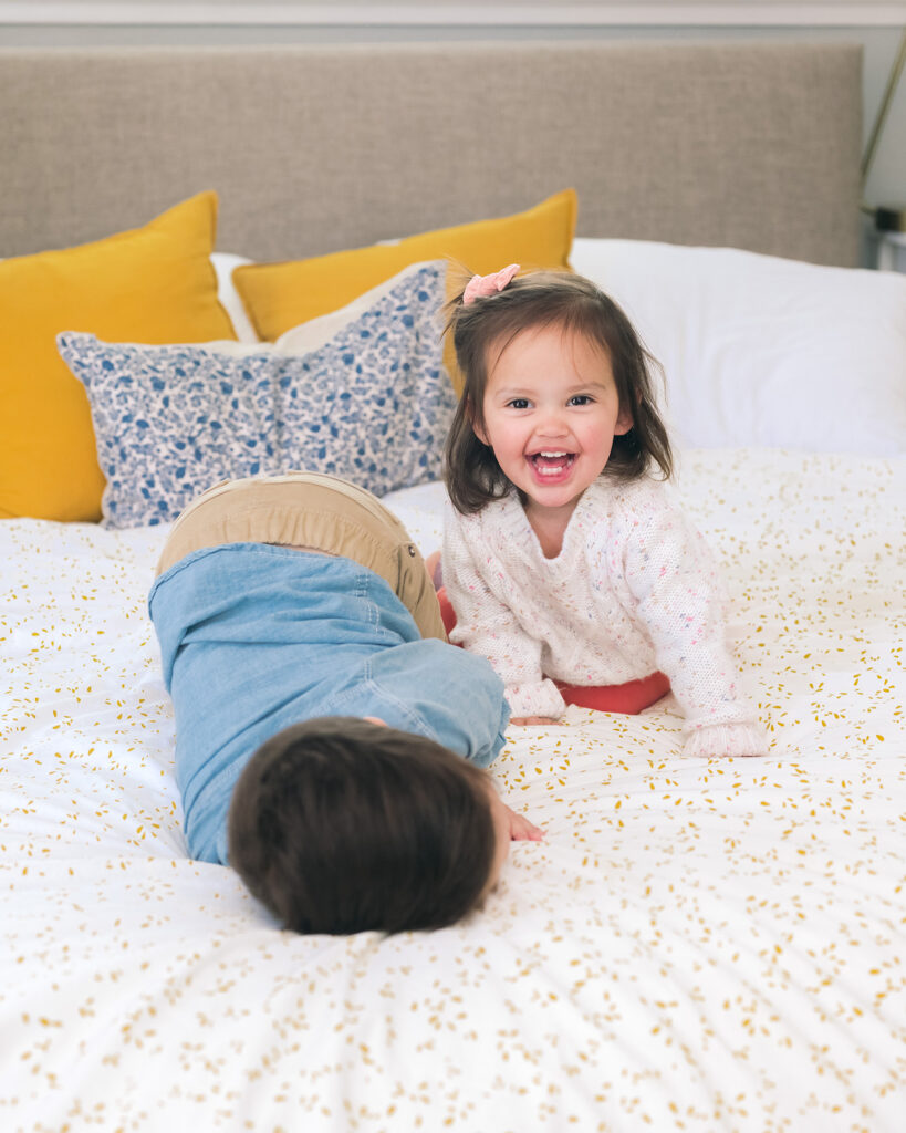 Toddler girl laughing on parents' bed while brother plays next to her during a relaxed family moment