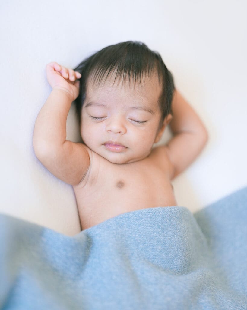 Close-up photo of a shirtless Indian baby boy sleeping on a white bed under a blue blanket, captured by a Palo Alto in-home newborn photographer using a Fujifilm GFX 50R camera.
