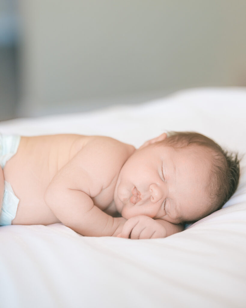 Close-up of a newborn baby sleeping in only a diaper on a white bed with soft natural light — captured by El Cerrito newborn photographer Isela Lozano.