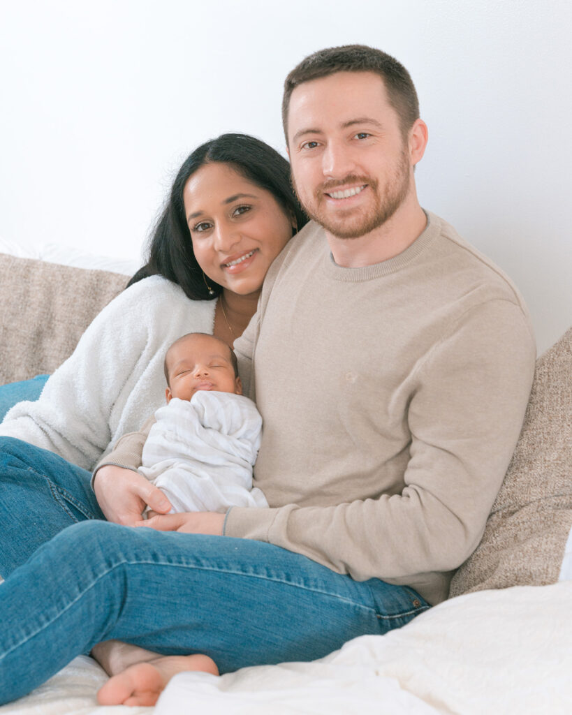 Mom and dad sitting on bed smiling at camera while holding their newborn baby boy swaddled in a blanket, El Cerrito newborn photography session.