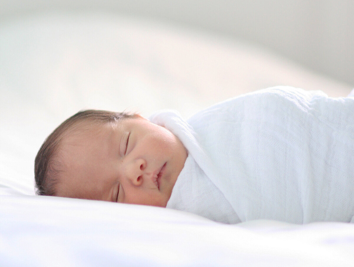 Newborn baby sleeping peacefully swaddled on a white bed in soft natural light — photographed by El Cerrito newborn photographer Isela Lozano.