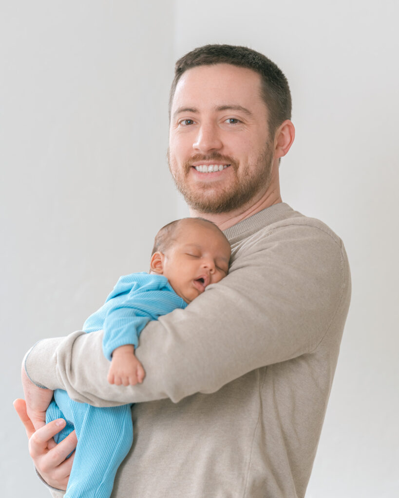 Dad holding newborn baby boy near a window, smiling at the camera, baby wearing a blue long sleeve onesie, El Cerrito newborn photography.