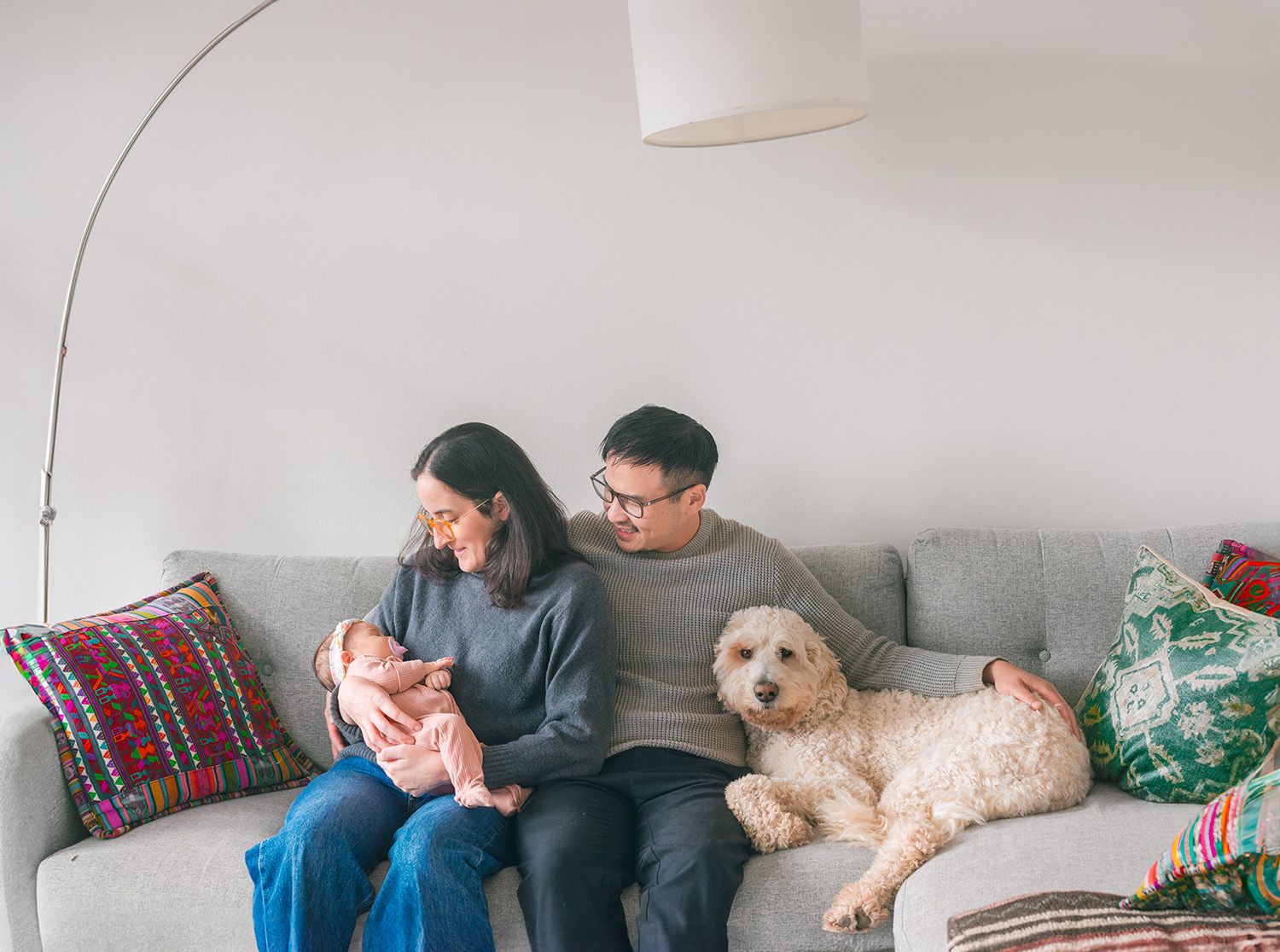 Mill Valley newborn photographer capturing an Asian father and white mother wearing glasses, sitting in their living room by a window, holding their newborn baby girl with their white labradoodle beside them during an in-home newborn session.