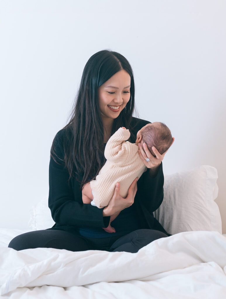 Mother sitting on bed holding her newborn baby in natural light.