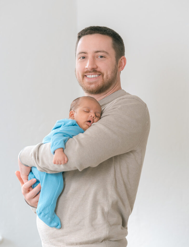 Sausalito newborn photographer capturing father in light brown sweater smiling at camera while holding newborn baby boy in blue long-sleeve onesie near window during in-home session.