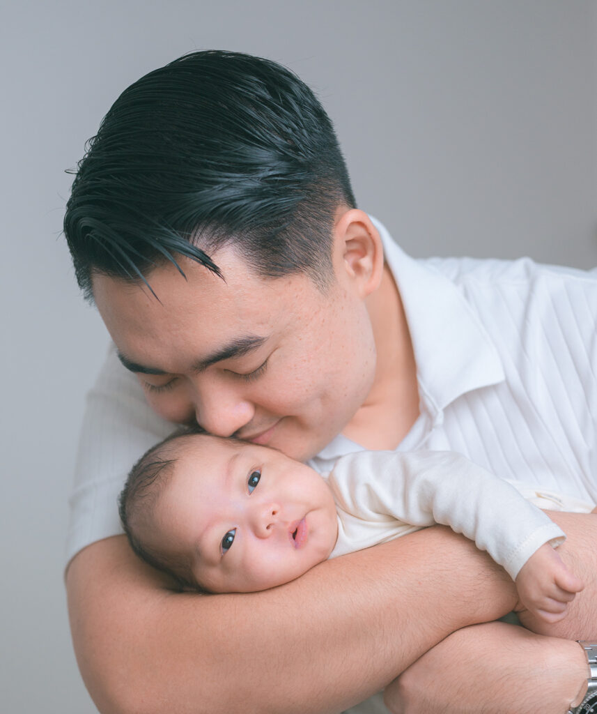 Father wearing white holding newborn baby in white onesie and kissing baby’s head.