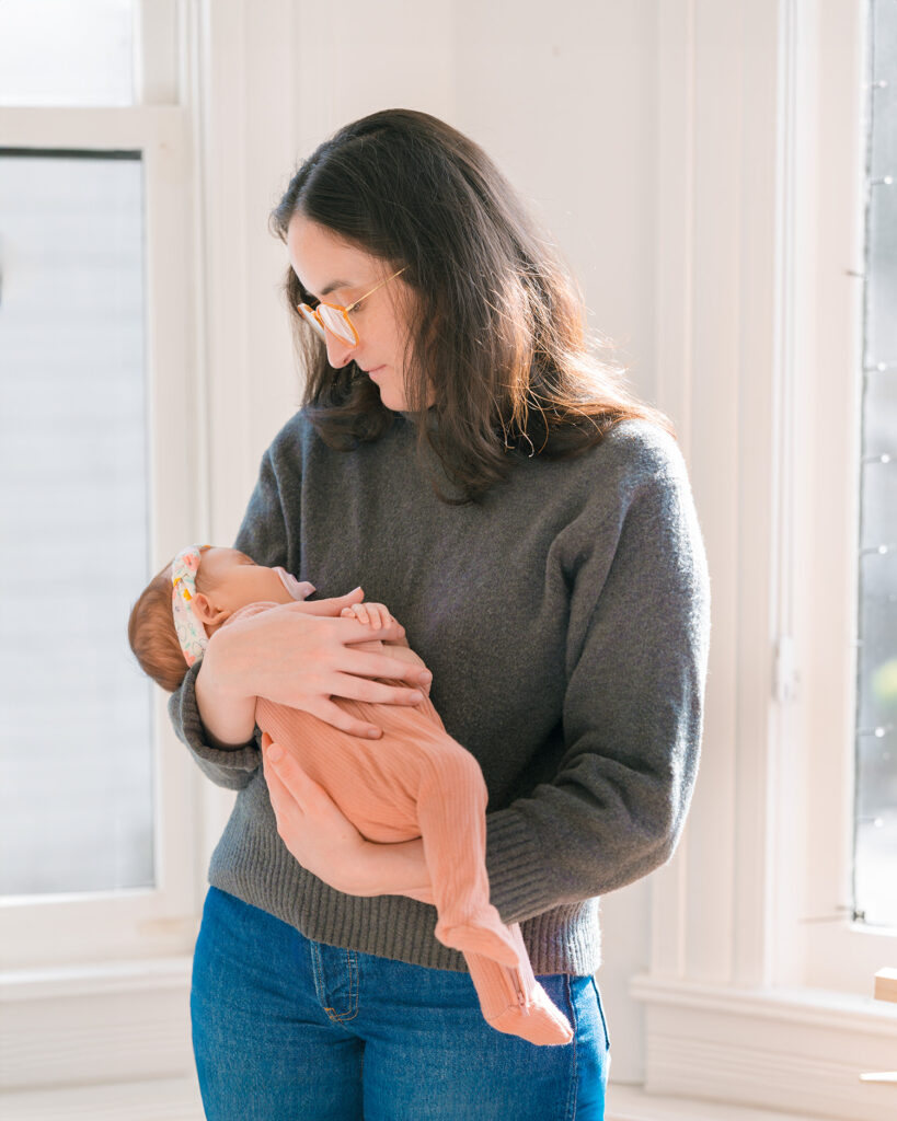 Palo Alto newborn photographer in-home session of a mother standing by a window holding her newborn baby girl wearing a pink long-sleeve onesie in her Palo Alto home.