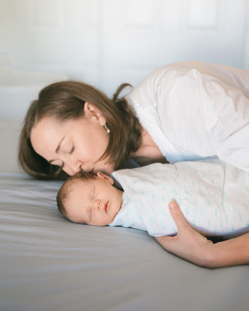 Newborn swaddled in a white blanket being kissed by mom on the bed during an in-home newborn photography session by Palo Alto photographer Isela Lozano.