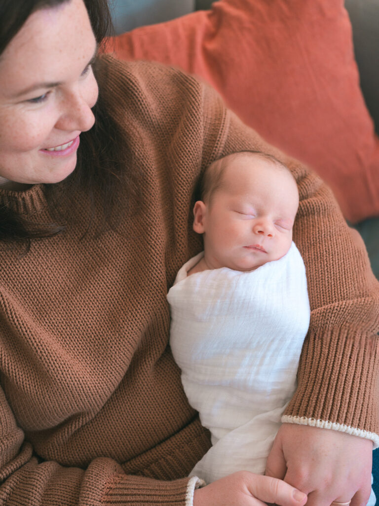 Palo Alto newborn photographer in-home session close-up of a mother wearing an orange long-sleeve sweater holding her sleeping newborn baby boy wrapped in a white swaddle blanket.