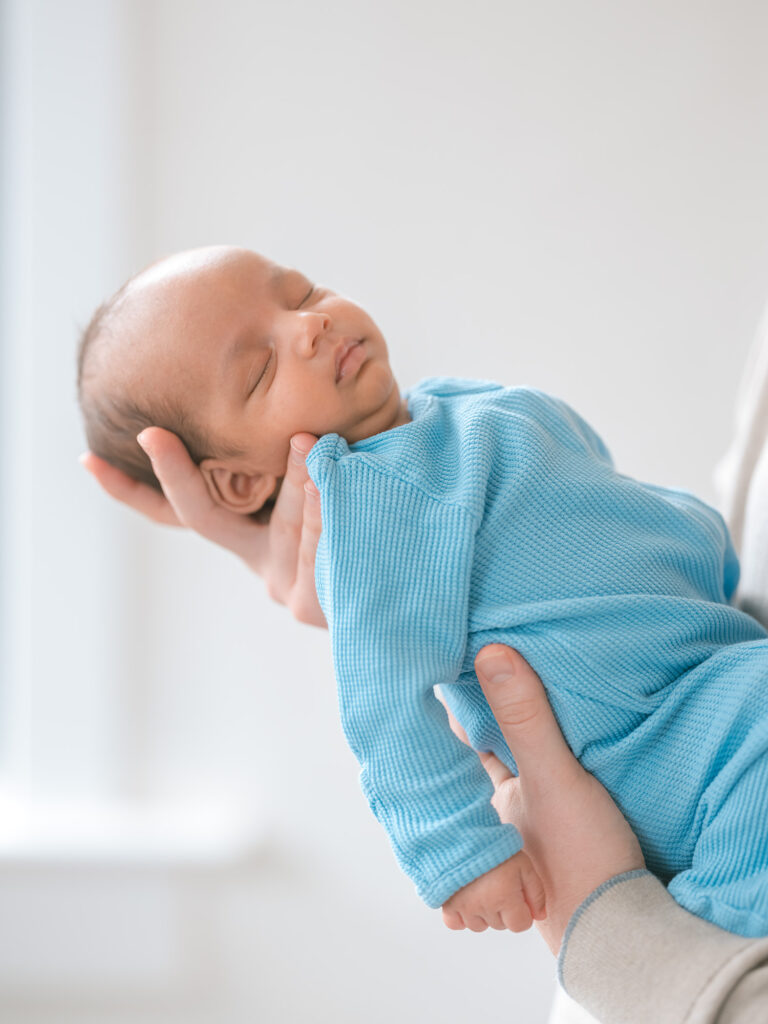 Palo Alto newborn photographer capturing close-up of newborn baby sleeping in blue long-sleeve onesie, gently held by father’s hands near window during in-home newborn photography session.