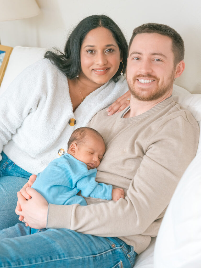 Parents sitting on white couch while father holds newborn baby in blue long-sleeve onesie sleeping on his stomach, both smiling at camera during in-home session, photographed by Palo Alto newborn photographer.