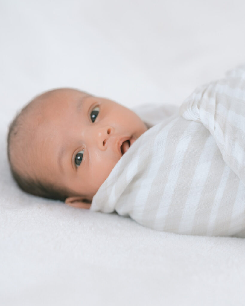 Swaddled newborn baby lying on bed looking at camera, captured in natural light by Palo Alto newborn photographer.