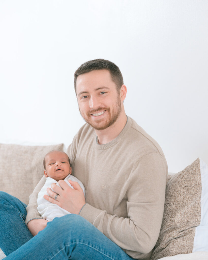 Father wearing jeans and light brown sweater holding newborn baby and smiling at camera on white bed with bright natural light, airy in-home portrait by Palo Alto newborn photographer.