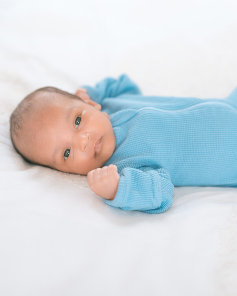 Close-up of newborn baby boy wearing blue long-sleeve onesie looking at camera while lying on white bed with natural window light during in-home newborn photography session in Sausalito.