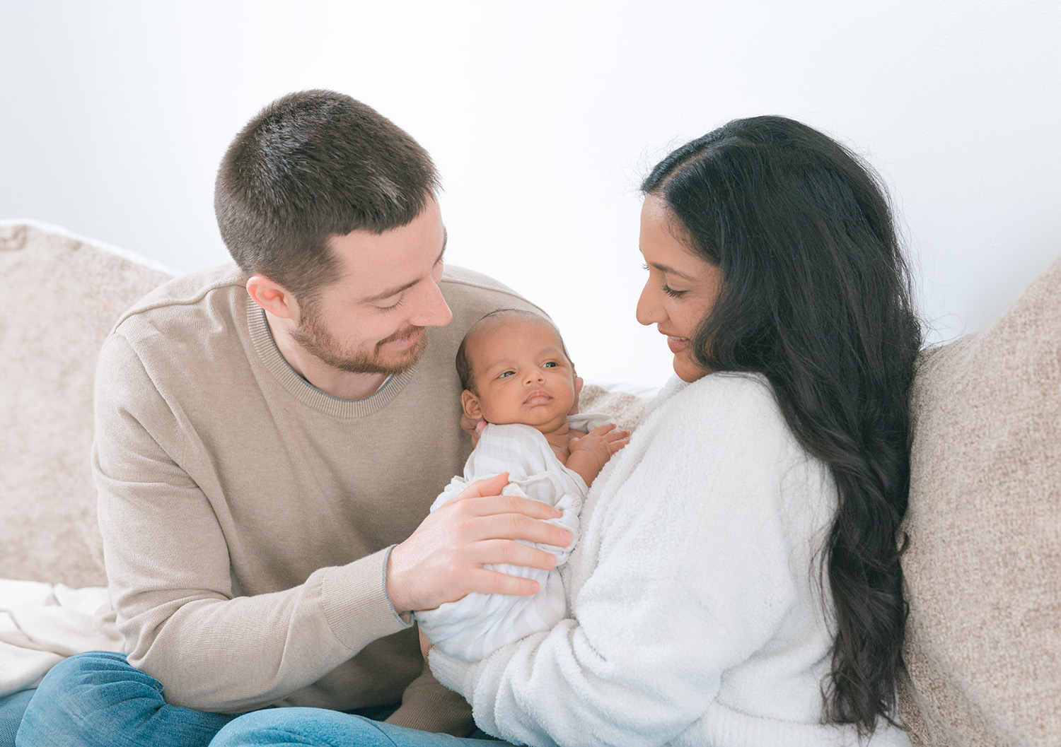 New parents sitting on a cozy beige couch at home in Sausalito, lovingly holding their newborn baby wrapped in a white swaddle during a natural light in-home newborn session by Sausalito newborn photographer Isela Lozano.