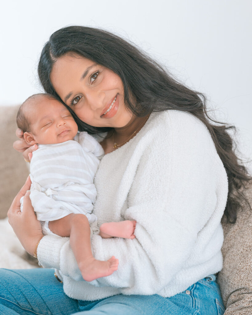 Sausalito newborn photographer capturing young Indian mother with long black hair in white sweater holding newborn baby close to her face, both smiling, during in-home newborn photography session.